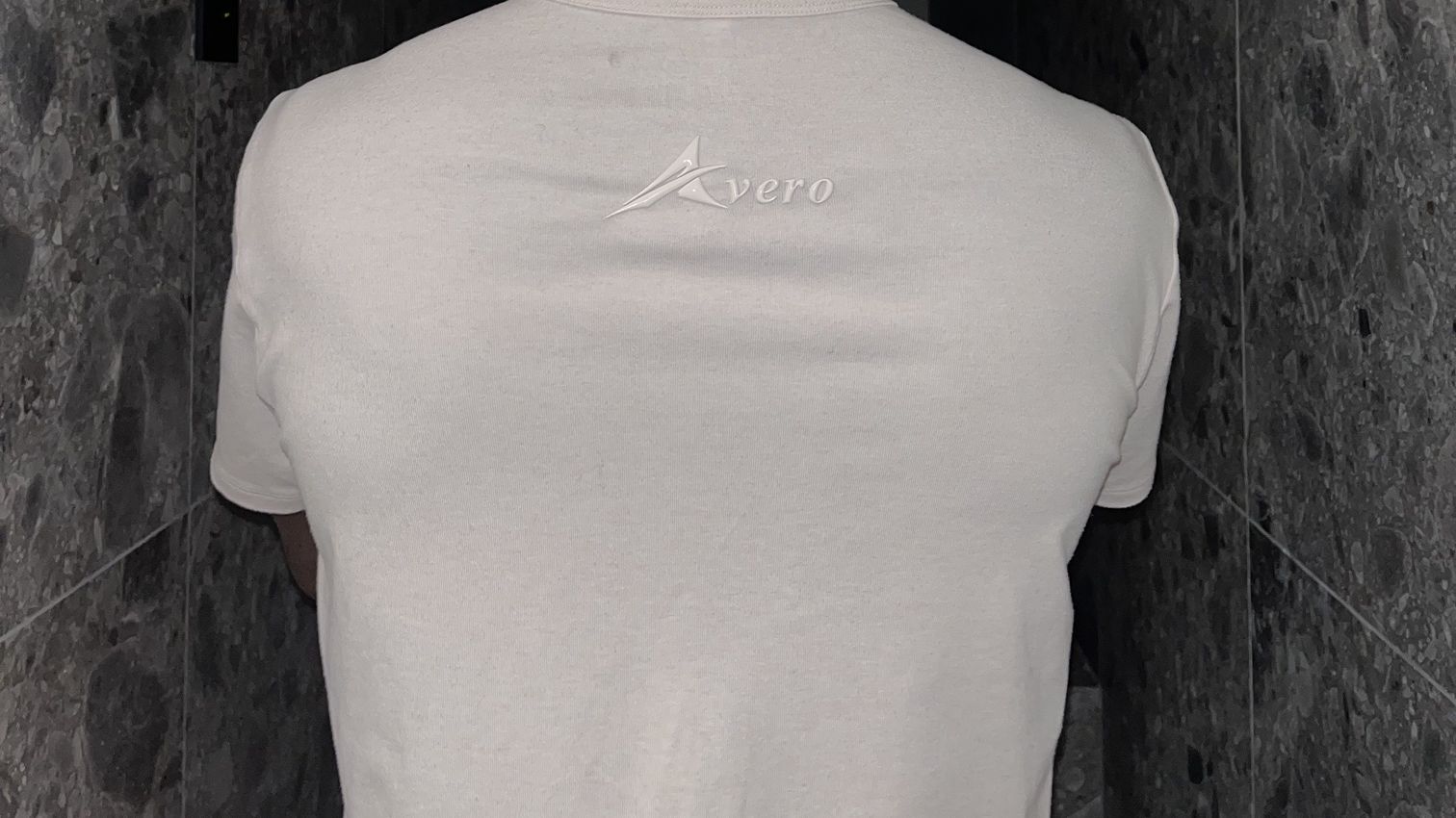 The back of the Avero Origin Tee in Sand, worn, with the tonal Avero logo below the collar